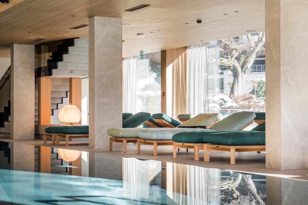 Mūsdienīgs interjers ar atpūtas krēsliem un lieliem logiem, kas nodrošina dabisku apgaismojumu. Redzama baseina atspulgs un eleganta kāpņu konstrukcija.