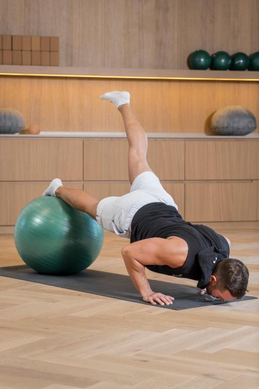Vīrietis veic vingrojumu uz sporta bumbas, balstoties uz vienas rokas un apgriežot kāju gaisā. Fons ir minimālisma stilā ar koka mēbelēm un dekoratīviem elementiem.