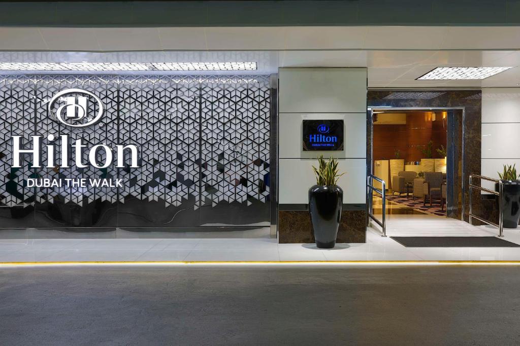 Hilton viesnīcas ieeja Dubaijā, ar modernu dizainu un stilīgu logo, kas uzsver viesnīcas atrašanās vietu pie "The Walk". Ieejas zona ir eleganta un aicina viesus iepazīt viesnīcas piedāvājumu.