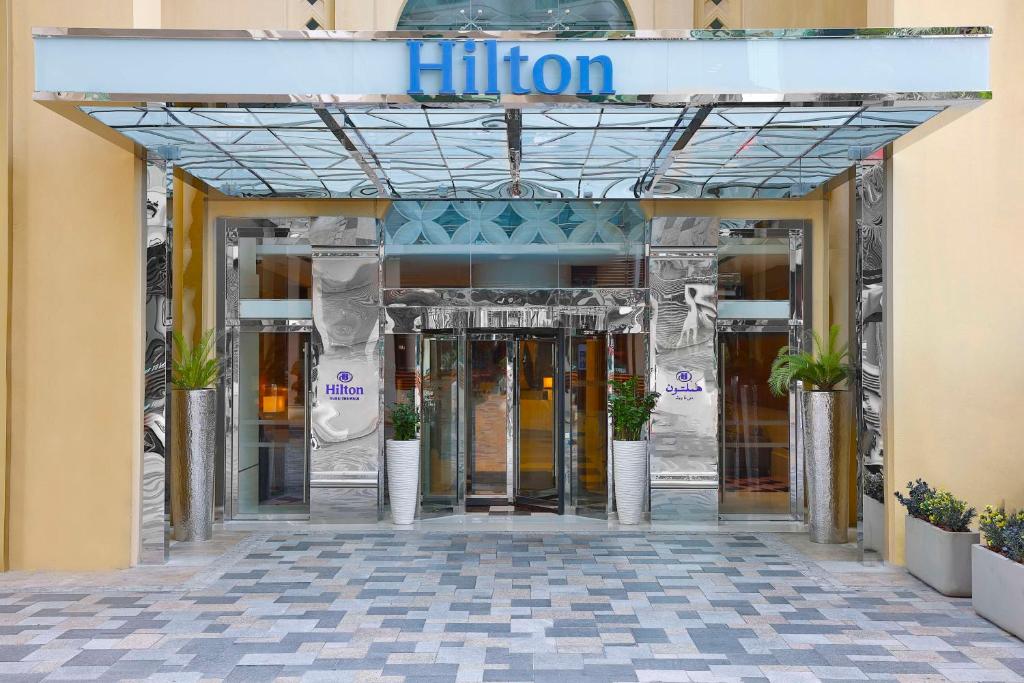 Hilton viesnīcas ieeja ar modernu dizainu, ko rotā spoguļi un zaļumi, radot elegantu atmosfēru. Izcila vieta ērtiem ceļojumiem un atpūtai.