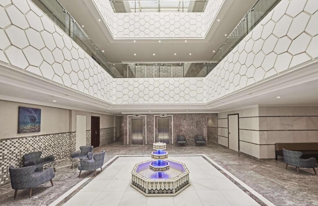 Mūsdienīga viesnīcas uzgaidāmā zona ar elegantu arhitektūru, hexagonālu sienu dizainu un centrālo strūklaku, kas rada relaksējošu atmosfēru.
