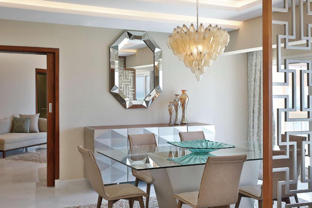 Mūsdienīgs ēdamistabas interjers ar stikla galdu, elegantu krēslu un dekoratīvu lustru. Uz sienas atrodas spogulis un dekoratīvi elementi, kas piešķir telpai eleganci.