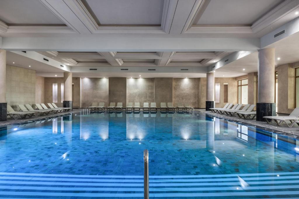 Mūsdienīgs iekštelpu peldbaseins ar gaišu interjeru un ērtām sauļošanās krēsliem ap to. Ūdens ir skaidrs un atspoguļo apkārtējo arhitektūru.