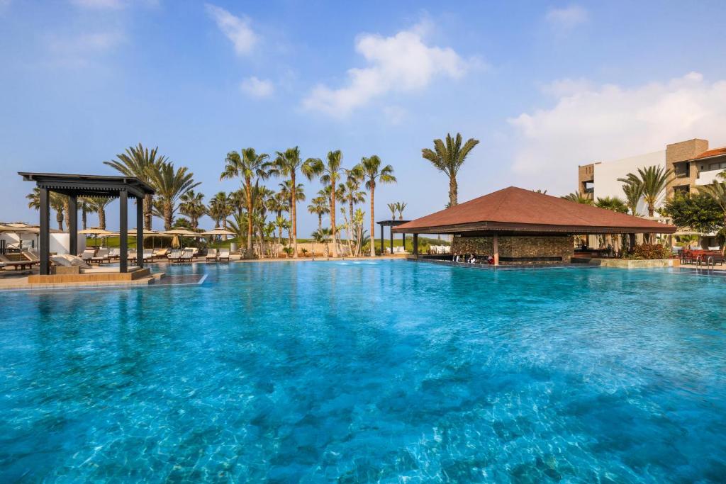 Luksusa viesnīcas baseins ar palmām un atpūtas zonām, kas piedāvā relaksējošu atmosfēru. Skats uz dzidru ūdeni un saulainām debesīm.