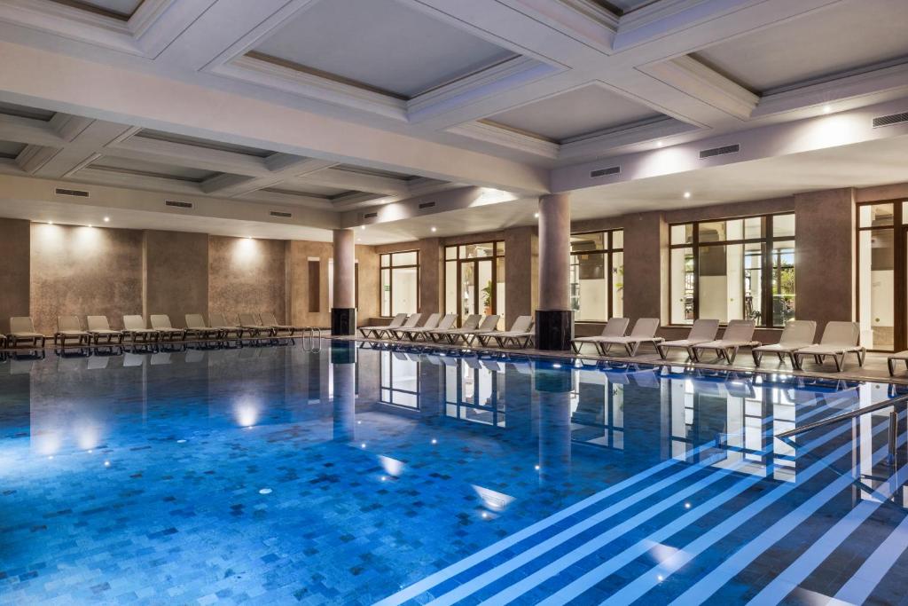 Luksusa iekštelpu peldbaseins ar zilu ūdeni un modernām saulessargu vietām, radot relaksējošu atmosfēru. Elegants dizains un apgaismojums piešķir vietai mierīgu noskaņu.