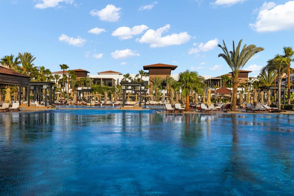 Luksusa viesnīcas peldbaseins ar palmām un atpūtas zonām, kas nodrošina relaksējošu atmosfēru. Debesis ir skaidras un zila, radot ideālu vidi brīvdienām.