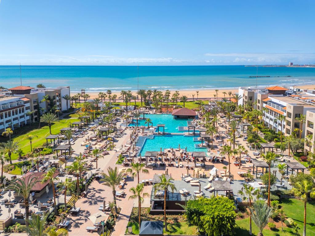 Viesnīcas kompleksa skats ar baseinu un pludmali, saule spīd un cilvēki atpūšas. Apkārt ir palmas un sauļošanās krēsli, radot atpūtas atmosfēru.