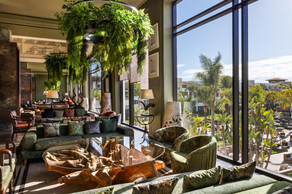 Mūsdienīga viesnīcas viesistaba ar greznām mēbelēm un lieliem logiem, caur kuriem redzams dārza skats. Interjerā ir zaļas augu kompozīcijas un dekoratīvi spilveni, radot relaksējošu atmosfēru.