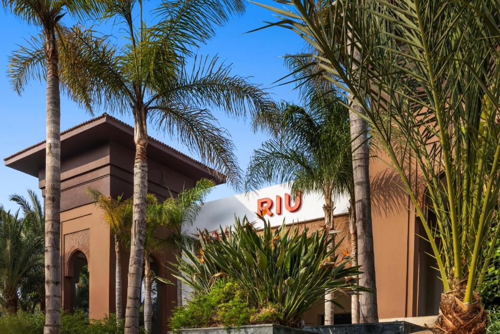 Riu viesnīcas ieeja ar palmām un zaļām augu kompozīcijām apkārt. Skats uz modernu arhitektūru un zilu debesu fonu.