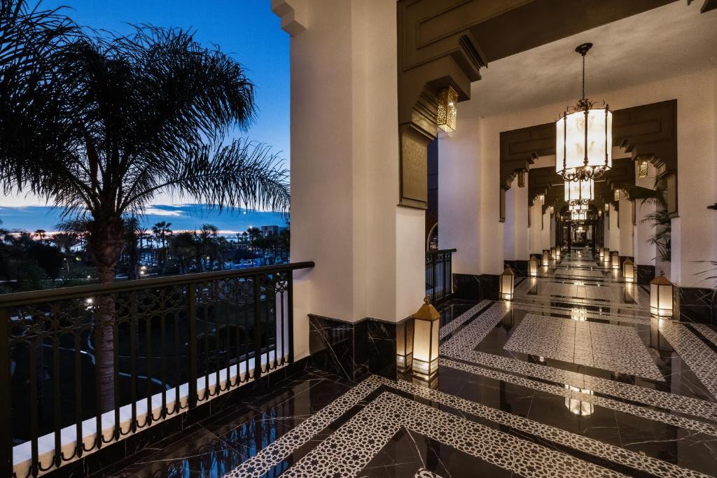 Eleganta gaitene ar melnā marmora grīdu un greznām lampām, kas rada romantisku atmosfēru vakara stundā. Aiz logiem redzama palmām apstādīta ainava pie jūras.