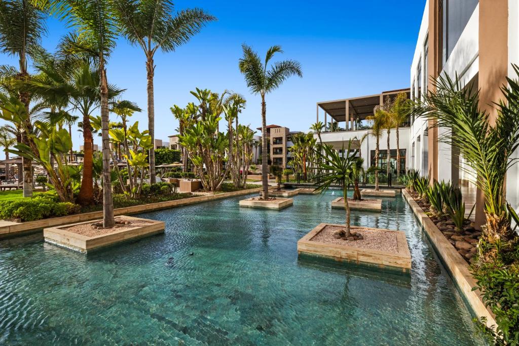 Tropu dārzs ar skaidru ūdeni un palmu stādījumiem, kas veido relaksējošu atmosfēru. Apkārt dārzam redzamas viesnīcas ēkas un saulains debesis.