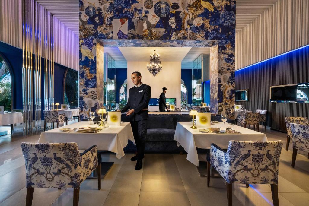 Elegantā restorāna interjers ar stilīgiem galdiem un apgaismojumu, kur apkalpo viesmīlis. Sienas rotā krāsains, māksliniecīgs tapetes dizains.