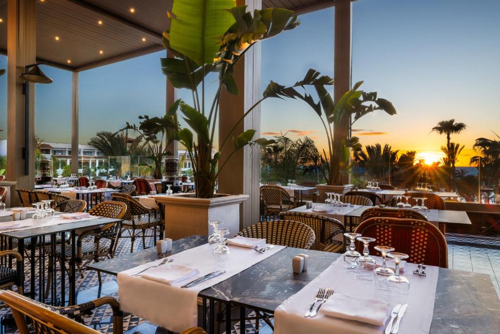 Moderna restorāna interjers ar ērtām sēdvietām, dekorēts ar augiem, kas piedāvā brīnišķīgu skatu uz saulrietu. Galdi ir klāti ar elegantiem galda piederumiem un glāzēm.