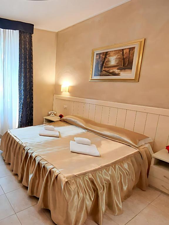 Mūsdienīgs viesnīcas numurs ar divguļamo gultu, kas rotāts ar zeltainu gultas veļu un baltām dvieļiem. Telpā ir eleganta apgaismošana un mākslas darbs uz sienas, radot komfortablu atmosfēru.