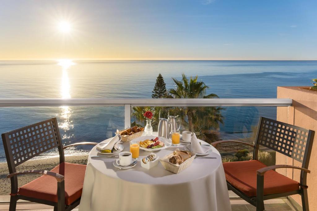 Brosta ar skatu uz jūru, kur uz galda atrodas brokastis un dzērieni, kamēr saule lec virs horizonta.