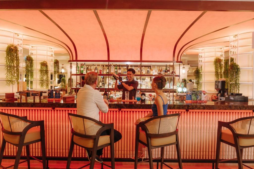 Moderns bārs ar stilīgu interjeru, kur bārmenis pasniedz kokteiļus diviem viesiem. Aiz bāra redzama plaša dzērienu izvēle un augu dekorācijas, radot patīkamu atmosfēru.