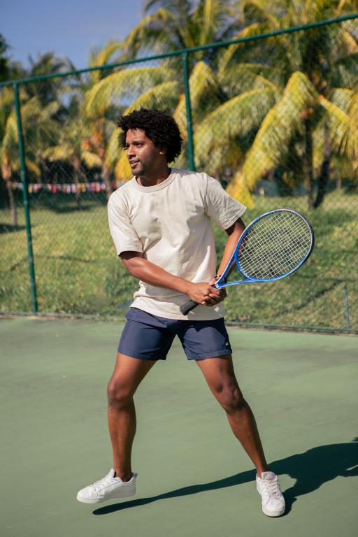 Vīrietis spēlē tenisu, turot raketi un gatavojoties izdarīt sitienu. Fons ir zaļa tenisa korta vide ar palmām.