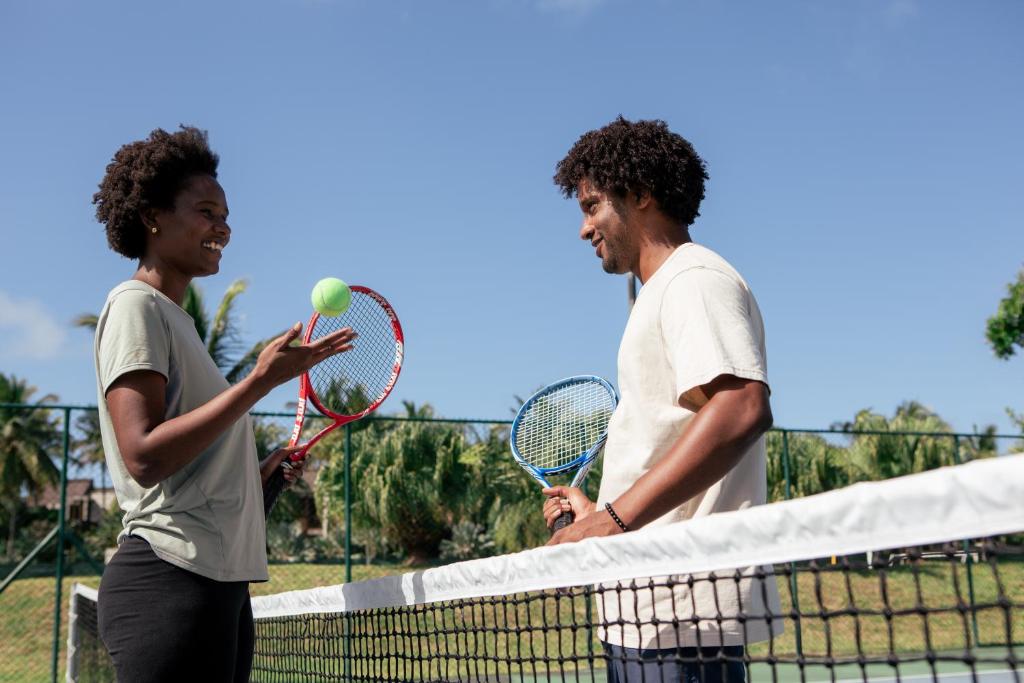 Divi cilvēki, kas spēlē tenisu, sarunājas pie tenisa laukuma, turēdami raketes un bumbu. Aiz viņiem redzami palmām apauguši apvidus un skaidras debesis.