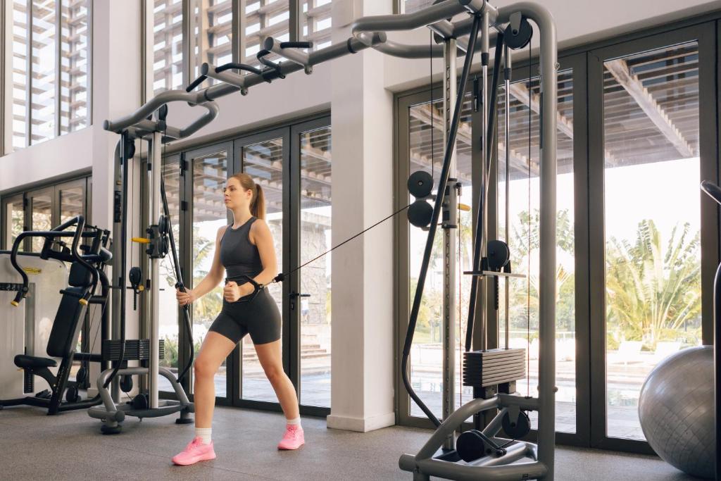 Sieviete sporto modernā sporta zālē, izmantojot trenažieri ar stieņu un svariem. Gaismas pilna telpa ar logiem un bagātīgu zaļumu fonā.