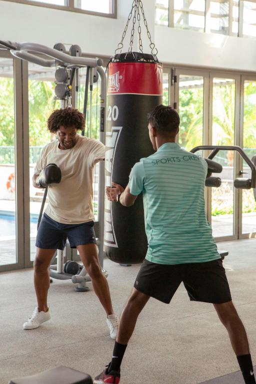 Divi vīrieši trenējas sporta zālē, sitot pa boksa maisu. Viens no viņiem ir apģērbts gaišā kreklā un šortos, otrs valkā sporta kreklu un šortus, abi koncentrējušies uz treniņu.
