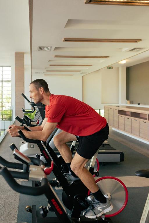 Vīrietis, kas trenējas sporta zālē uz velotrenažiera, koncentrējoties uz savu vingrinājumu. Gaismas, kas ieplūst telpā, rada motivējošu atmosfēru.