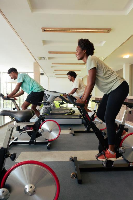 Vingrošanas zālē trīs cilvēki trenējas uz velotrenažieriem, aktīvi piedaloties grupas nodarbībā. Gaisma ir dabiska, un telpa ir moderna un plaša.