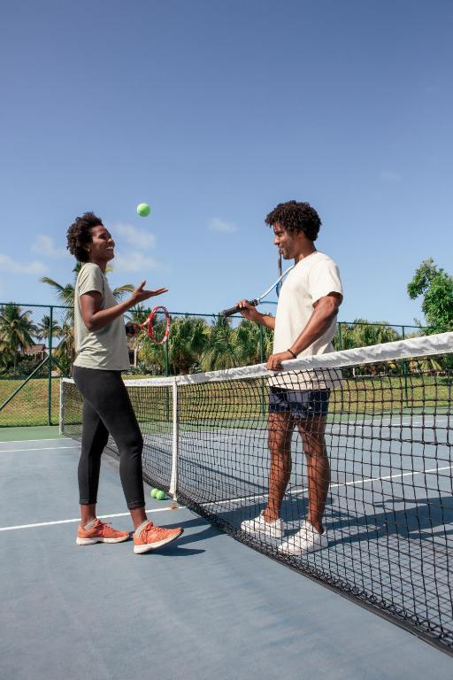 Divi cilvēki sarunājas tenisā, sieviete met bumbiņu gaisā, kamēr vīrietis stāv pie tīkla ar raketi. Apkārt ir zaļi koki un skaidrs debesis.