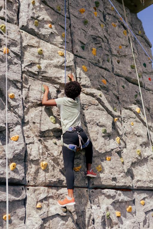 Kāpējs, kas cenšas sasniegt virsotni mākslīgajā klinšu kāpšanas sienā, izmantojot dažādus turēšanas punktus. Sienā ir vairākas krāsainas rokas turēšanas, kas palīdz kāpšanas procesā.