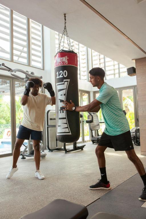 Divi vīrieši trenējas sporta zālē, viens no viņiem sit pa boksa maisu, kamēr otrs sniedz instrukcijas. Apkārt ir sporta iekārtas un lieli logi, caur kuriem ieplūst dabiska gaisma.