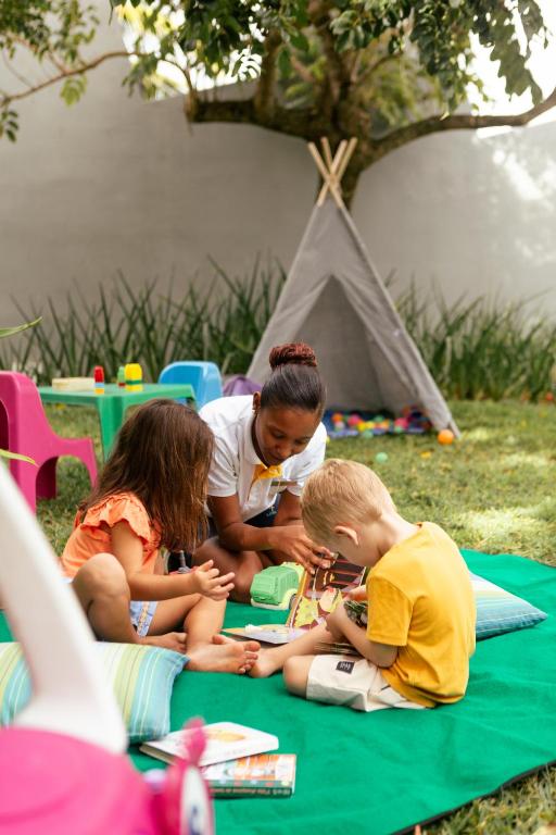 Bērni spēlējas dārzā, izmantojot radošas rotaļlietas, kamēr pieaugušais viņiem palīdz. Fons satur telti un krāsainas rotaļlietas.
