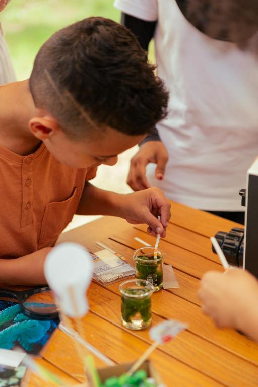 Bērns veic eksperimentu ar zaļām vielām, izmantojot stikla traukus un koka lāpstiņu. Apkārt ir citi bērni un dažādi eksperimenta rīki.