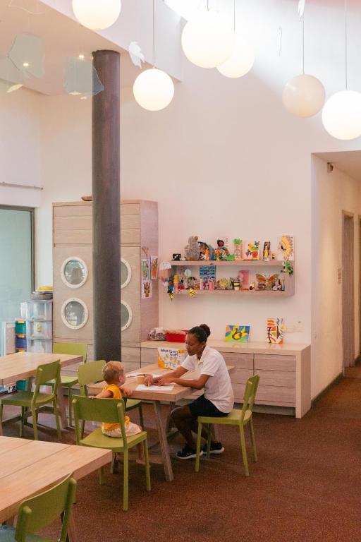 Gaiša un draudzīga bērnu telpa, kur pie galda māca bērnu un pieaugušo. Uz plaukta redzami dažādi mākslas darbi un rotaļlietas.