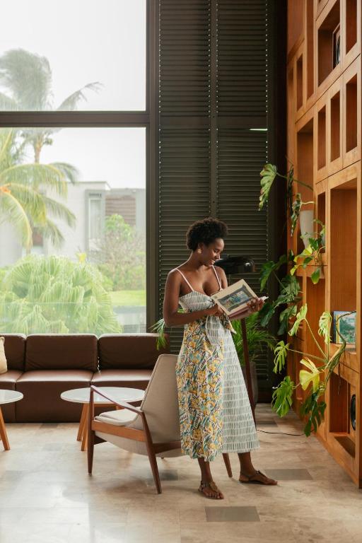 Sieviete ar īsiem matiem lasa grāmatu mūsdienīgā viesistabā ar lieliem logiem un zaļiem augiem. Interjers ir gaišs un elegants, ar dīvānu un koka plauktiem.