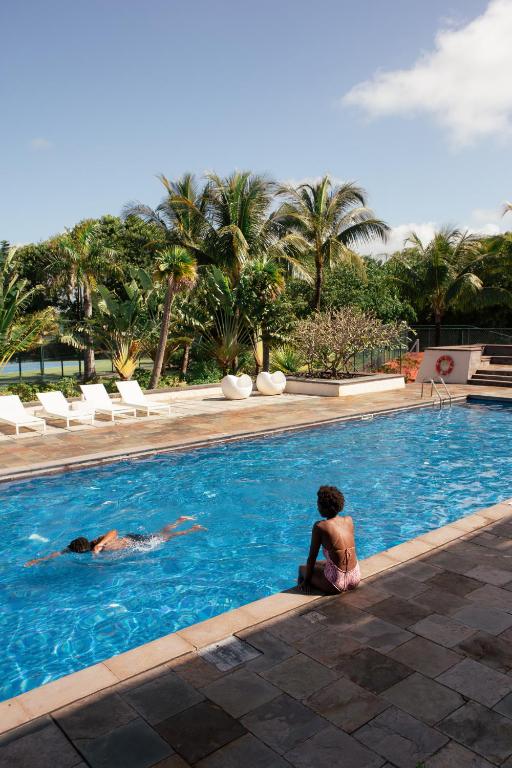 Peldbaseins ar palmām un atpūtas zonu, kur divi cilvēki izbauda sauli un ūdeni. Ideāla vieta relaksācijai un atpūtai.