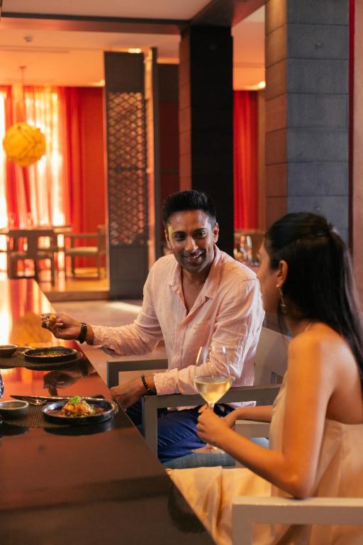 Divi cilvēki, vīrietis un sieviete, sēž pie galda modernā restorānā, baudot maltīti un sarunājoties. Aiz viņiem ir elegants interjers ar sarkaniem aizkariem un apgaismojumu.