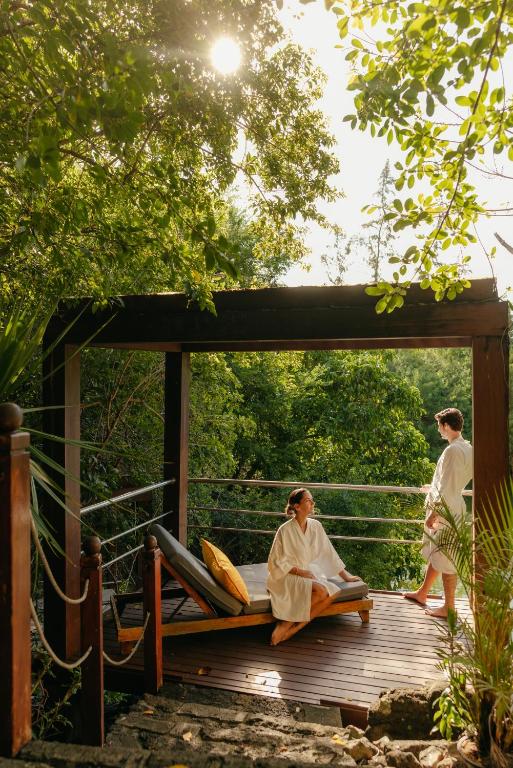 Relaksējoša atmosfēra spa zonā, kur divi cilvēki ģērbušies baltos halātos, izbauda dabu un mieru. Apkārt ir zaļi koki un saules gaisma, kas caurspīdina lapotnes.