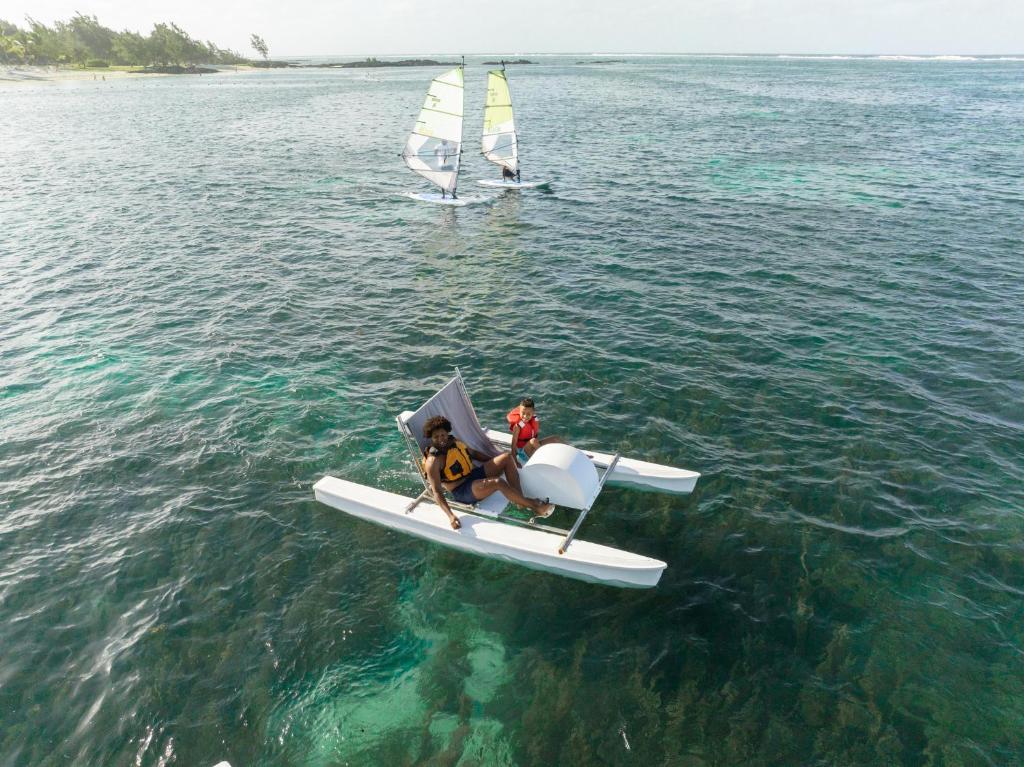 Divi cilvēki brauc ar katamarānu skaidrā ūdenī, kamēr fona pludināšanas laivas izpilda sportiskas aktivitātes. Skats uz jūru ar zilo ūdeni un piekrasti.