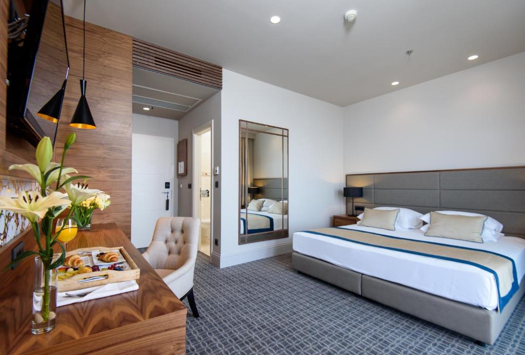 Mūsdienīgs viesnīcas numurs ar ērtu divguļamo gultu, elegantu mēbelējumu un dekoratīvām detaļām, piemēram, ziediem un brokastu komplektu uz galda. Gaismas un spoguļa elementi piešķir telpai plašuma sajūtu.