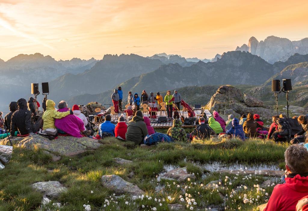 Mūzikas pasākums kalnos ar skatītājiem, kas sēž uz zālāja, un izpildītājiem uz skatuves. Aiz muguras redzami kalnu ainavas un saulriets.