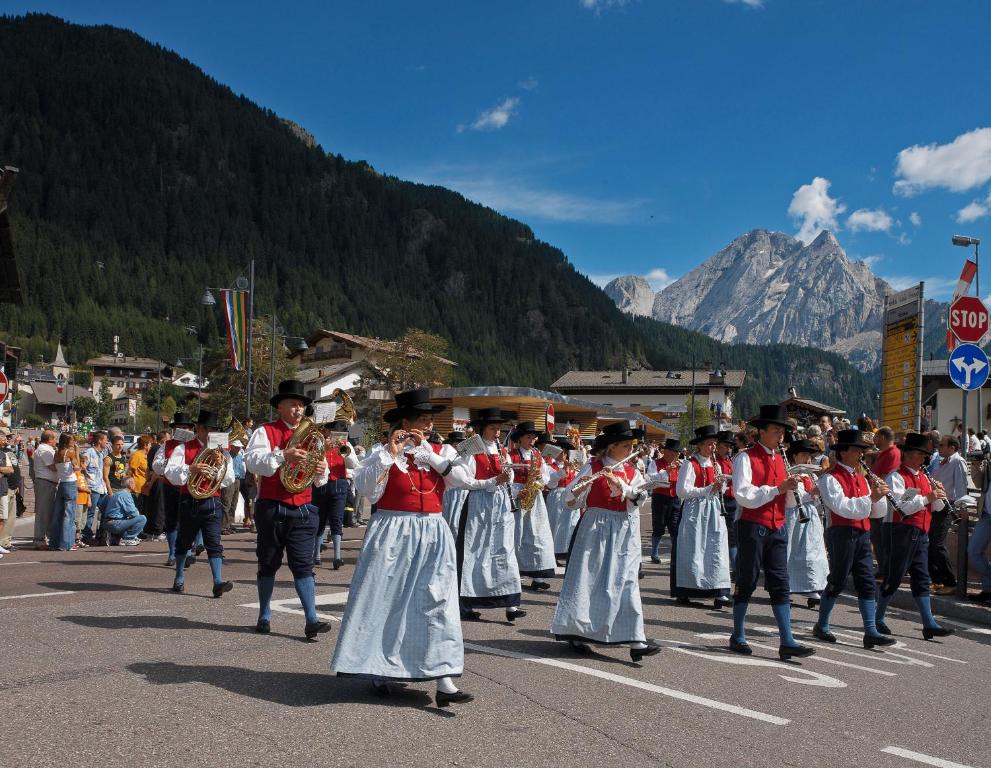 Tradicionāla gājiena skats ar muzicējošiem dalībniekiem, tērptiem tradicionālā apģērbā, kalnu ainavā. Pasākums notiek apdzīvotā vietā, kur redzami arī skatītāji un ceļa zīmes.