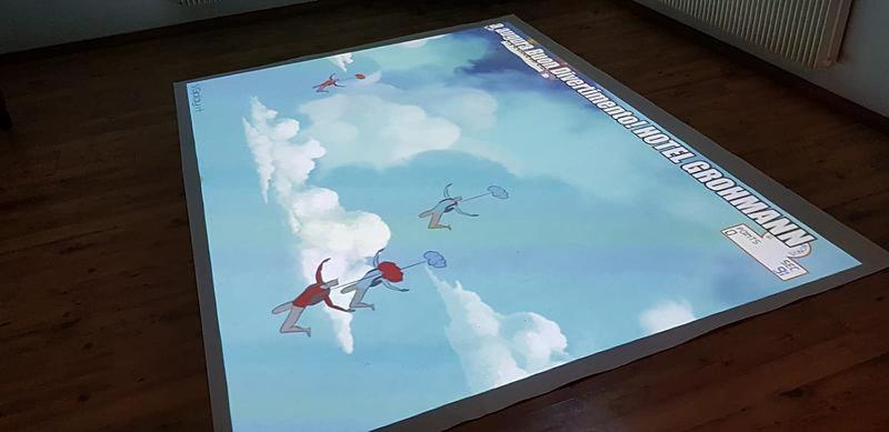 Grīdas projekcija ar zilu debesu fonu, kurā redzami stilizēti cilvēki, kas lido ar baloniem. Projekcija satur arī tekstu, kas saistīts ar viesnīcu "GROHMANN".