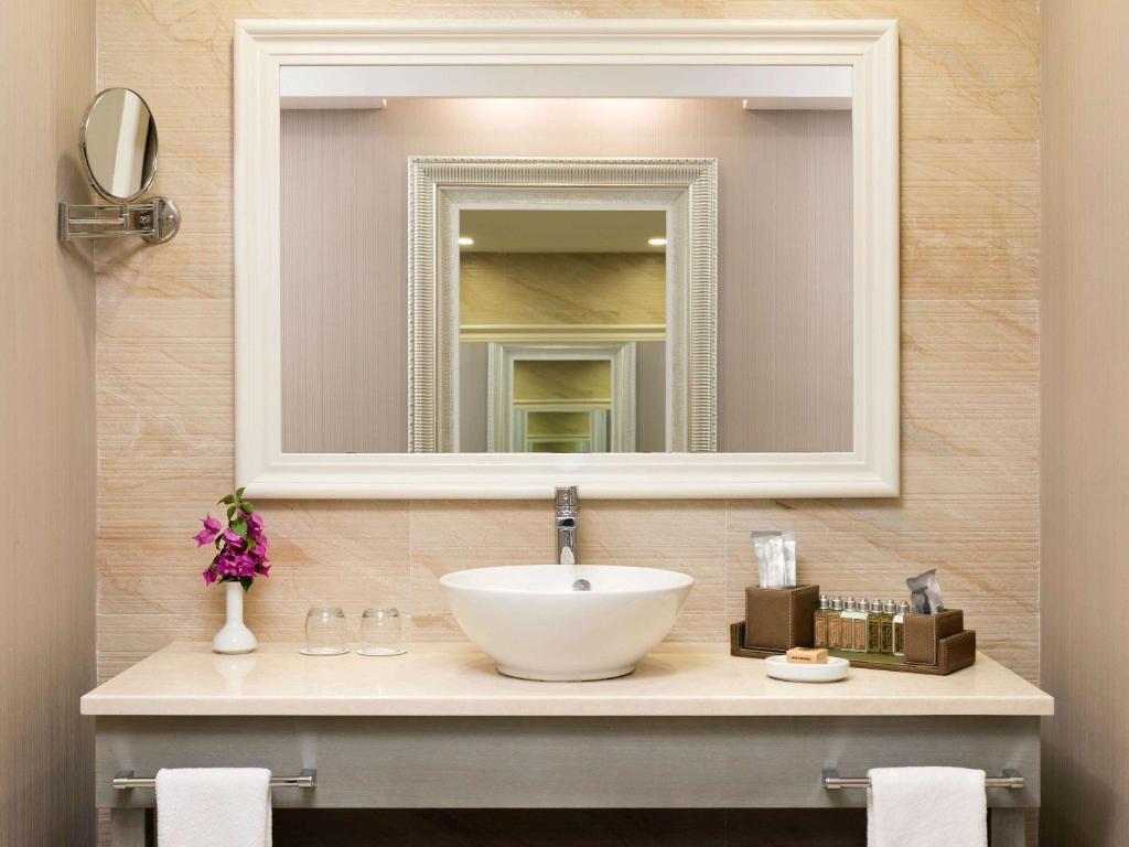 Mūsdienīgs vannas istabas interjers ar baltu izlietni, spoguli un dekoratīvām detaļām, tostarp violetām ziedu kompozīcijām un higiēnas piederumiem.