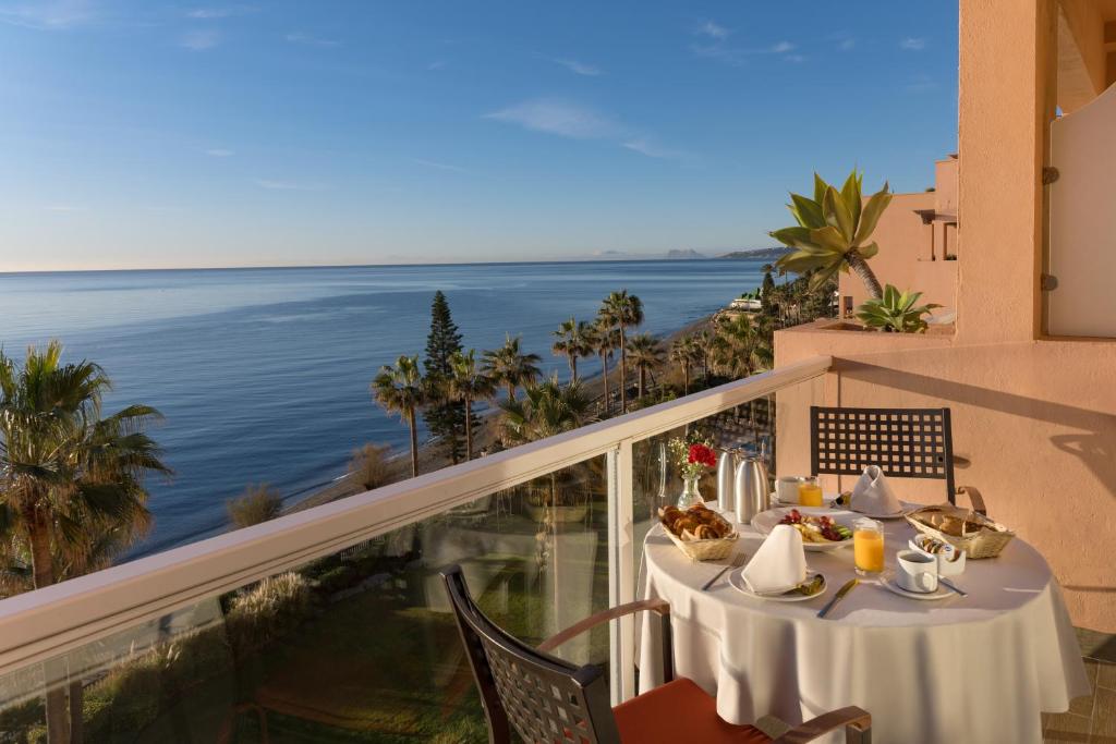 Brokastis ar skatu uz jūru no balkona, kur uz galda ir dažādi ēdieni un dzērieni, ieskauj palmām un saulainu laiku.