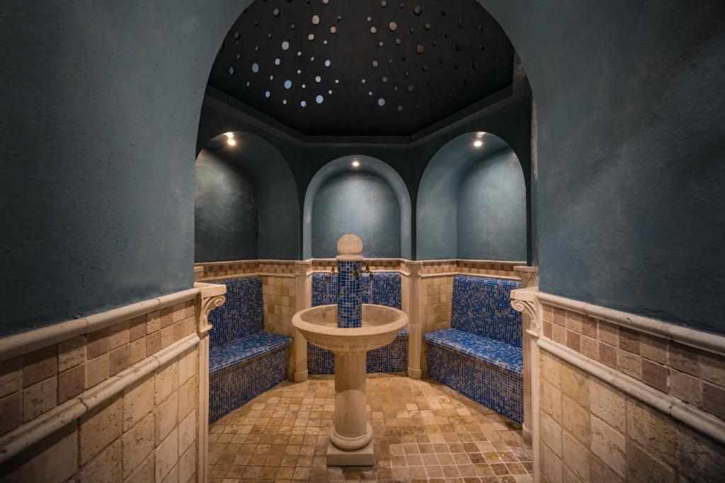 Eleganta tvaika pirts ar zilu mozaīku un arku nišām, kur centrā atrodas dekoratīva strūklaka. Telpa ir apgaismota ar mīkstu gaismu, radot relaksējošu atmosfēru.