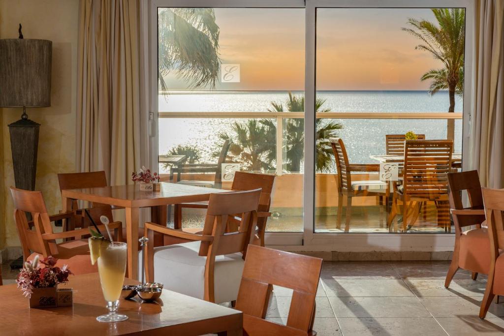 Elegants restorāns ar skatu uz jūru, kurā redzami koka galdi un krēsli, kā arī viesmīlīga atmosfēra. Logā redzamas palmas un saulriets, kas rada relaksējošu noskaņu.