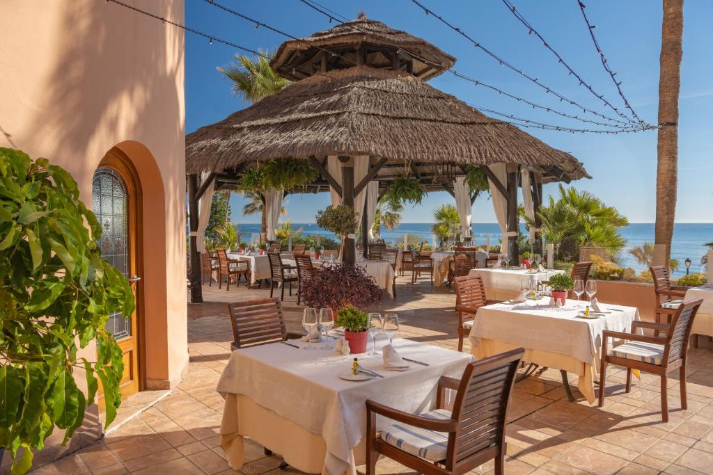 Āra restorāna zona ar salmu jumtu, elegantām galdiem un krēsliem, kas vērsti uz jūru, apzaļumotu vidi un saulainu laiku. Ideāla vieta relaksācijai un maltītei.