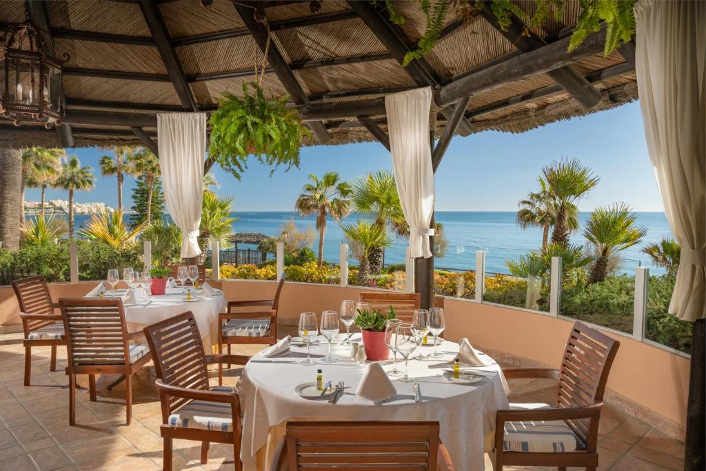 Elegantā terase ar skatu uz jūru, kur uz galdiem ir sakārtoti trauki un glāzes. Apkārt ir tropu augi un saulains laiks, radot relaksējošu atmosfēru.