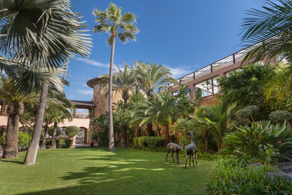 Skaists dārza skats ar palmām un zaļo zāli, kurā novietotas divas statujas, kas attēlo briežus. Aiz dārza var redzēt modernu ēku ar stikla elementiem.