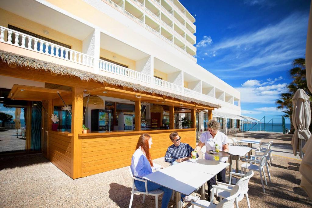 Āra kafejnīca pie pludmales ar viesmīli, kas apkalpo viesus pie galdiem, saulains debesis un jūra fonā. Ideāla vieta atpūtai un baudīšanai.