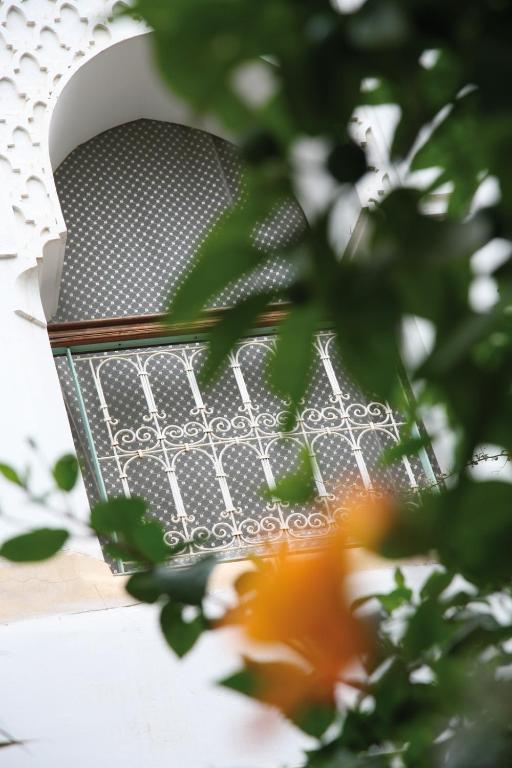 Alogu raksts par arābu arhitektūru, kurā redzams loga fragments ar dekoratīvu metāla resti un rakstainu aizmuguri, ko ieskauj zaļumi un ziedi.